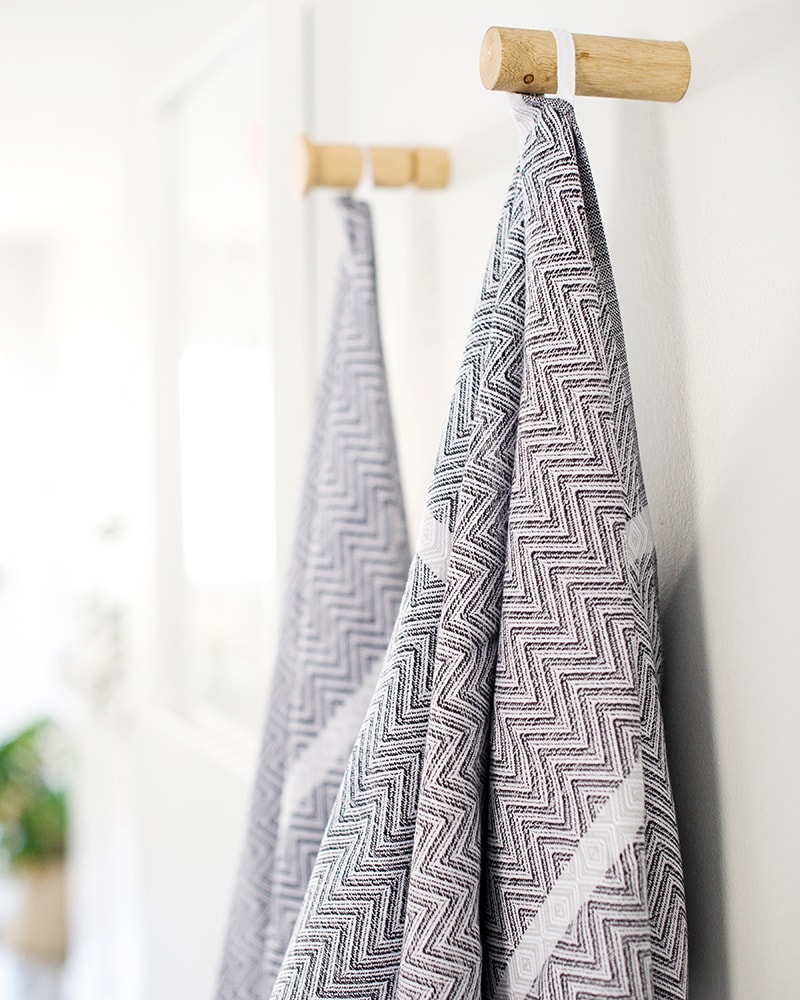 Mungo Tawulo Cotton Towels in Thunder Grey, hanging on pegs in bathroom scene. Textile woven from all natural fibres at our mill in Plettenberg Bay.