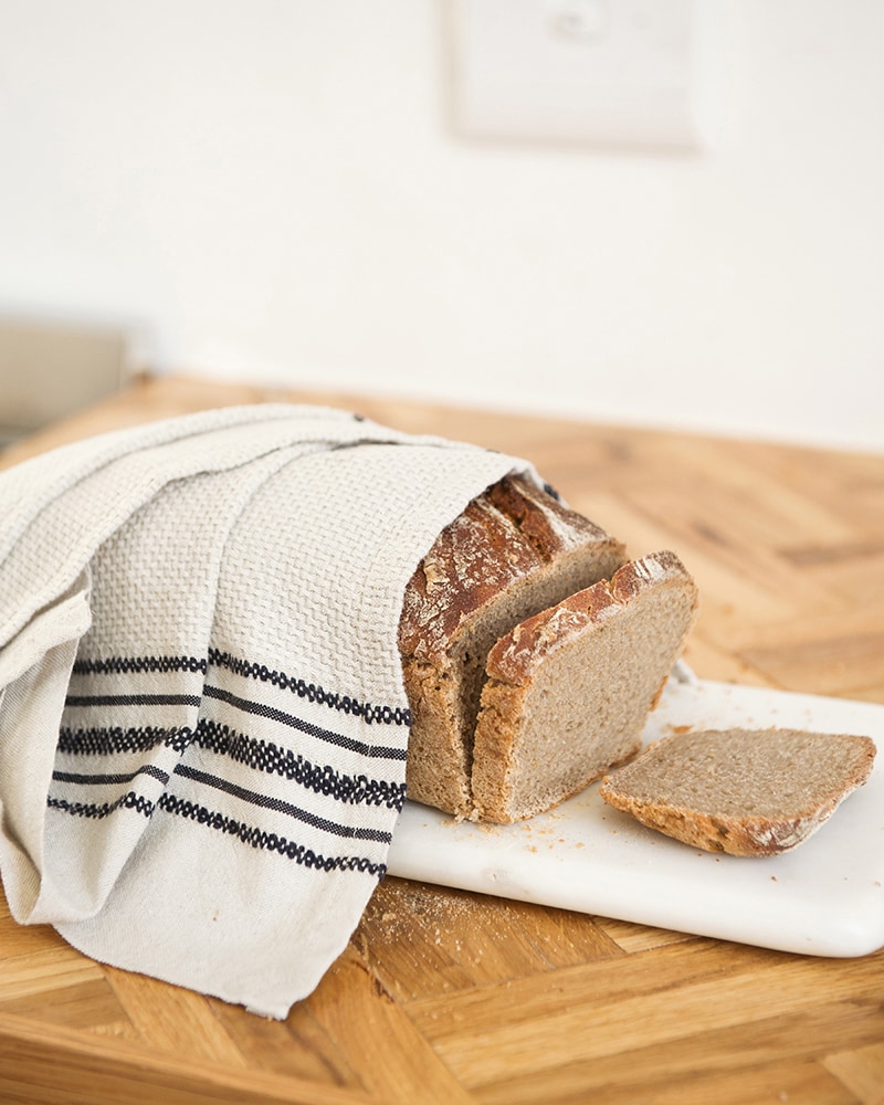 A Mungo Linen Kitchen Utility Cloth in Natural, on a wooden country table with fresh bread.