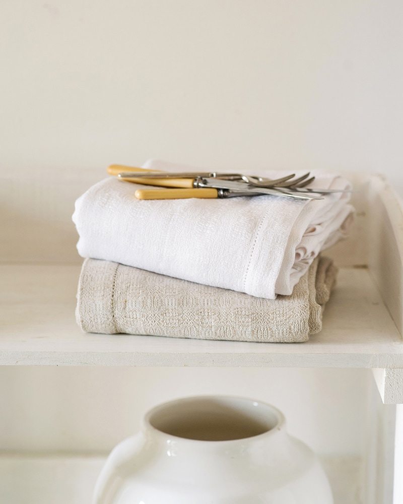 The Mungo Cloverleaf Linen Table Cloth folded in Natural Flax and White on the shelf of a country kitchen.